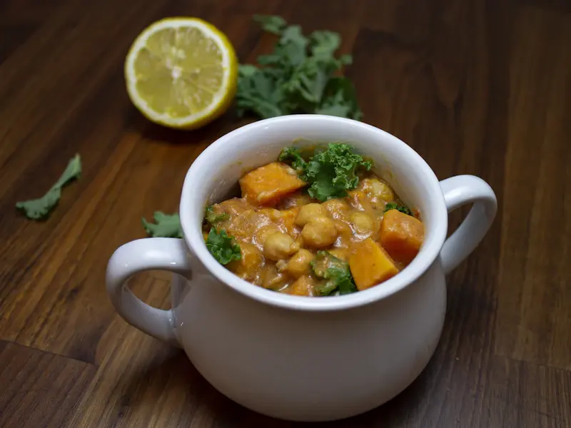 Una ciotola bianca di curry di ceci con zucca e cavolo nero, con mezzo limone su un tavolo di legno