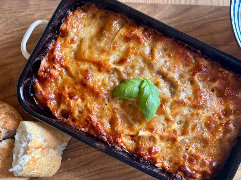 Una teglia di lasagna dorata e fumante con una fogliolina di basilico, accanto a del pane croccante